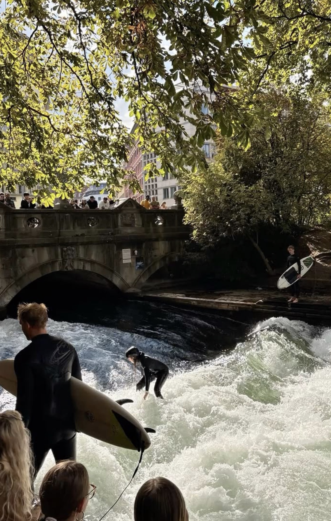 People surfing a standing river wave under a stone bridge